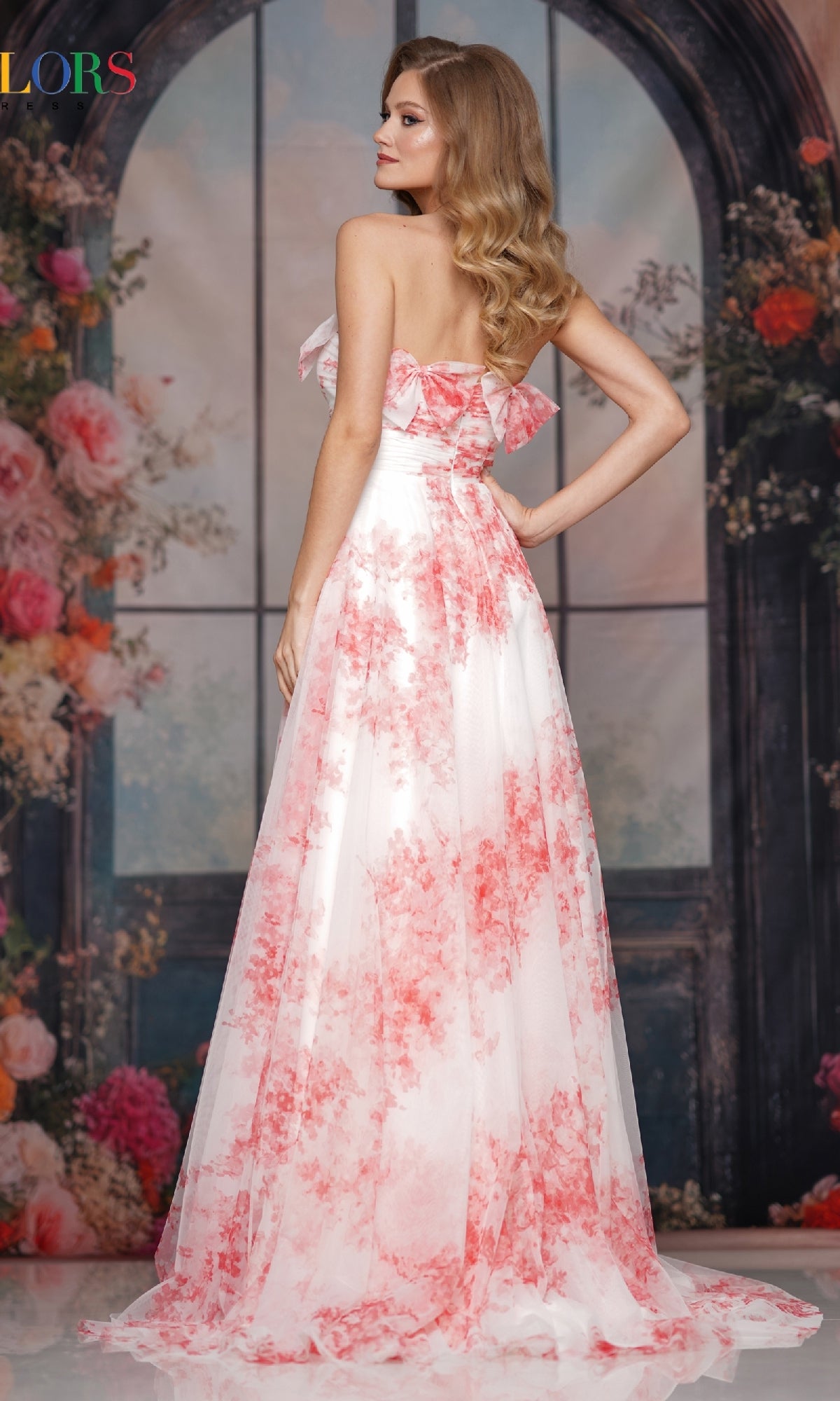 A woman with wavy blonde hair faces away, wearing the Colors Dress 3811—an A-line, strapless long prom dress with pink floral patterns—posed before a floral and arched window backdrop.